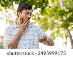 Serious dissatisfied Indian man stands outdoors looking for someone waiting for meeting angry frustrated looks time pointing finger at wristwatch feeling upset about being late lateness. Horizontal