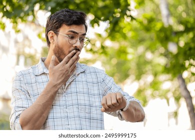 Serious dissatisfied Indian man stands outdoors looking for someone waiting for meeting angry frustrated looks time pointing finger at wristwatch feeling upset about being late lateness. Horizontal