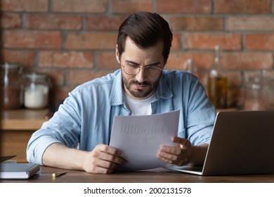 Serious distance employee analyzing statistics, data in paper financial reports at workplace with laptop. Focused young man reading document, loan agreement with payment schedule. Paperwork concept