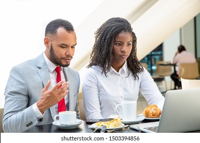 Serious Diverse Office Colleagues Watching Presentation On Laptop. Business Man And Woman Sitting In Cafe, Drinking Coffee, Eating, Looking At Computer Screen. Coffee Break Concept