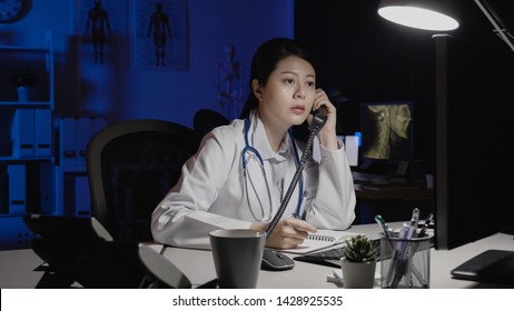 Serious Doctor Answer Phone Call Working In Clinic Office At Late Night. Young Female Nurse Discussing With Medical Surgeon About Patient Situation Staring At Computer Screen In Midnight On Telephone