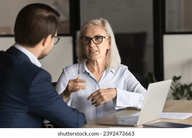 Serious elder professional woman talking to younger colleague man at workplace, working on project together, discussing collaboration, management strategy. Business partners speaking at office table - Powered by Shutterstock