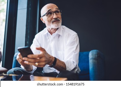 Serious elderly 60 years old businessman in glasses sitting in comfy chair near bright window, holding mobile phone in hands and looking away during break - Powered by Shutterstock