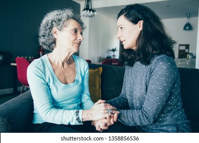 Serious Elderly Woman And Her Daughter Talking And Holding Hands. Mother And Daughter Sitting On Couch With Home Interior In Background. Daughter And Elderly Mother Concept.