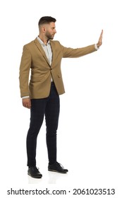 Serious Elegant Handsome Man In Beige Jacket Is Standing With Hand Raised And Showing Stop Gesture. Side View. Full Length Studio Shot Isolated On White