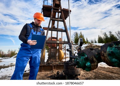 Serious Engineer In Work Overalls And Helmet Holding Clipboard And Looking At Oil Pump Rocker-machine. Worker Checking Oil Pumping Unit, Making Notes. Concept Of Oil Extraction And Petroleum Industry