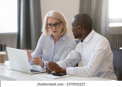 Serious experienced aged businesswoman in glasses explains help to african new employee task usage of corporate online program sitting at table office room working together, mentor and trainee concept