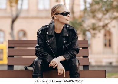 Serious fashionable woman with black sunglasses on her eyes looking away wearing a black t-shirt, black leather jacket and black leather pants sitting on a bench with her hands crossed on her knees