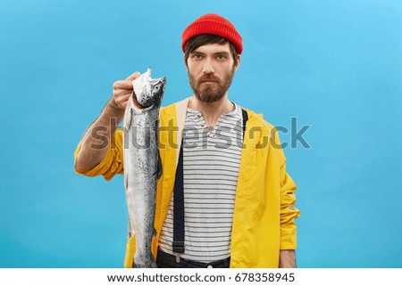 Serious fashoinable man with beard dressed in yellow raincoat and red hat holding big fish in hands. Handsome fisherman showing his huge catch during his vacations isolated over blue background