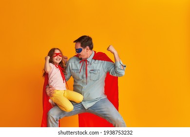 serious father with a beard, a superhero holding a little baby girl, together dressed in superman costumes, preparing for a children's party, isolated on a yellow wall. Strong heroes. copy-space