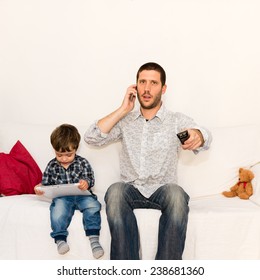 Serious Father Making A Phone Call And Watching Tv While Son Is Playing With Tablet On A White Sofa With A Red Pillow And A Bear Toy