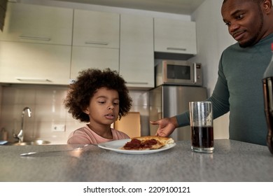 Serious Father Near Little Son Having Lunch Or Dinner With Cola And Pizza At Table At Home Kitchen. Unhealthy Eating. Young Black Family Lifestyle And Relationship. Fatherhood And Parenting