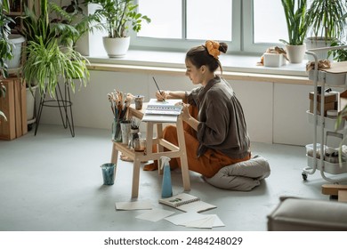 Serious female artist sitting on floor, painting with watercolor in sketchbook. Surrounded by professional art supplies tools unsmiling woman focused on drawing sketch with aquarelle, creative process - Powered by Shutterstock