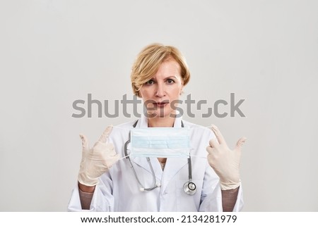 Serious female doctor holding and showing medical mask. Mature caucasian woman with stethoscope wearing white coat and latex gloves. Medical and health care. White background in studio. Copy space