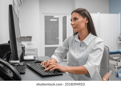 Serious female doctor using computer and writing notes in medical electronic journal sitting at desk. Young woman professional medic physician wearing white coat working on pc computer at workplace. - Powered by Shutterstock