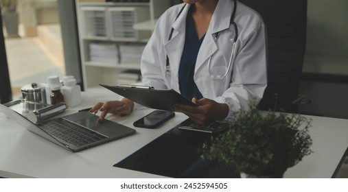 Serious female doctor using laptop and writing notes in medical journal sitting at desk. Young woman professional medic physician wearing white coat and stethoscope working on computer at workplace. - Powered by Shutterstock