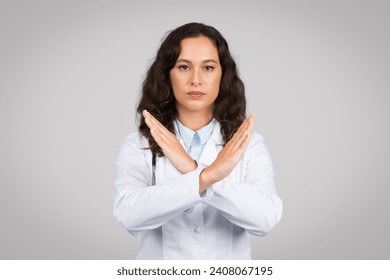 Serious female doctor in white coat making X sign with her arms, signaling denial or prohibition, wearing white lab coat with stethoscope, grey background