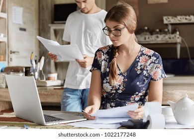 Serious female dressed in blouse, looks attentively at screen of laptop computer, holds documents, count expenses and taxes, prepares financil report or manages family budget together with husband