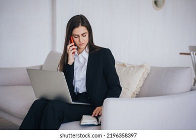 Serious Female Entrepreneur In Black Suit Talking By Phone Sitting On Couch With Laptop At Home Opens Her Diary Checking Agenda. Pretty Hispanic Upset Woman Received Bad News. Business And Finance.