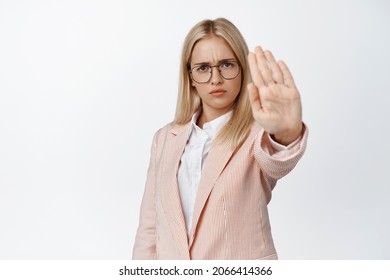 Serious female entrepreneur stretching out hand in stop sign, prohibit, forbid something, saying no, standing over white background in suit and glasses