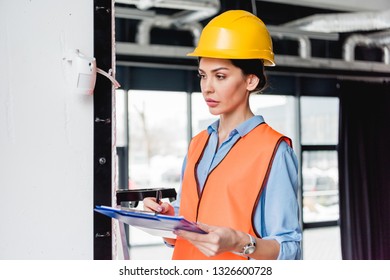 Serious Female Firefighter Standing Near Fire Alarm While Holding Clipboard