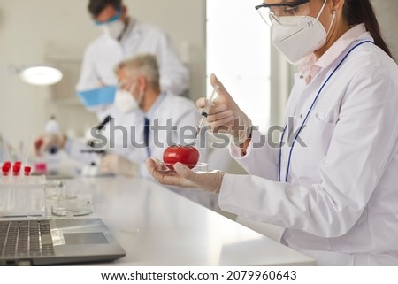 Serious female genetic scientist in white coat, protective goggles and face mask doing innovative research in food laboratory, developing GMO or pesticides and injecting tomato with chemical additive