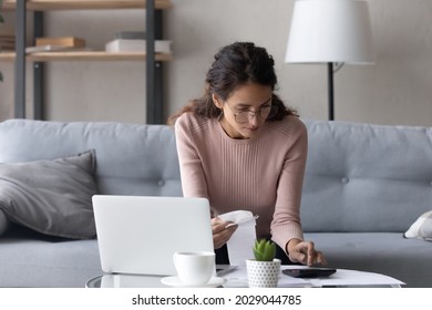 Serious female in glasses paying utility bills, taxes, rental fees use online services e-bank app on laptop. Young woman work from home sit on couch hold paper invoice calculate expenses, make report