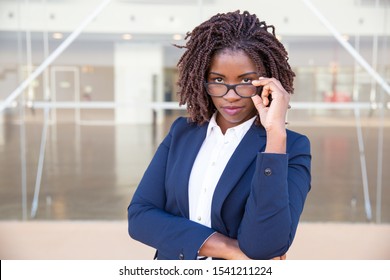 Serious Female Office Employee Touching Eyewear Outside. Young African American Business Woman Standing At Glass Wall, Adjusting Glasses, Looking At Camera. Vision Concept