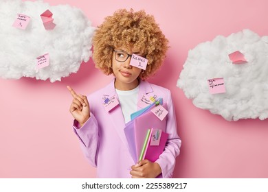 Serious female office worker coveres with memo stickers writes down list to do poses with folders indicates index finger overhead dressed in formal clothes isolated over pink background clouds above - Shutterstock ID 2255389217