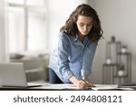 Serious female student, focused employee making notes on paper, writing plan, leans over desk, surrounded by papers and documents, deeply engrossed in workflow, review project details. Goals, tasks