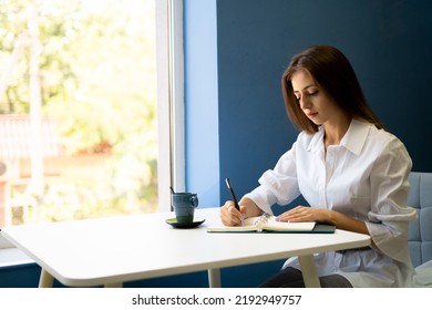 Serious female student writes a story in a notebook while sitting in a cafe with a cup of cappuccino. Focused young woman notes text in notepad