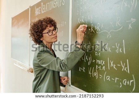 Serious female teacher of algebra with piece of chalk writing down equations on blackboard while explaining new subject