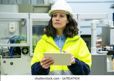 Serious Female Technician Holding Tablet. Confident Factory Worker Using Tablet While Standing Near Printing Machine. Print Manufacturing, Technology Concept