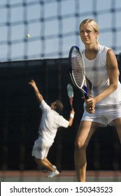 Serious female tennis player holding racket with partner serving ball in background