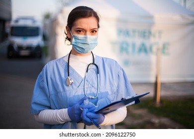 Serious Female UK NHS Doctor Wearing Protective Equipment Face Mask,standing Next To Triage Tent And Ambulance Car In Front Of Emergency Hospital,worried Stressed Look On Face,COVID-19 Pandemic Crisis