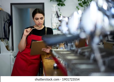 Serious female waitress in apron cafe uniform reading information from report checking accounting at bar during working process, pensive woman checking commercial audition organize job