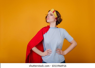 Serious Female Warrior In Funny Costume Posing On Yellow Background. Indoor Photo Of Curly Girl In Toy Crown And Red Cloak.