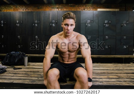 serious fitness model of a bodybuilder male trainer sitting on a bench in the locker room. after or before training. On the background of a bag, a phone and a bottle. Torso, muscles, six pack abs.