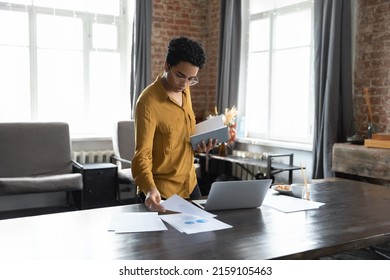 Serious Focused African Business Woman Sorting Out Papers, Analyze Financial Charts And Diagrams Shown On Report Standing Near Desk With Laptop. Doing Paperwork, Workflow In Office, Planning Concept