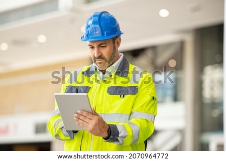 Serious and focused architect or engineer working on construction site using digital tablet. Mature man working on digital tablet at construction site. Mid adult manual worker checking list on laptop.