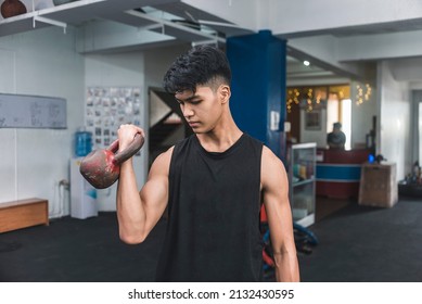 A Serious And Focused Asian Man In A Black Tank Top Working Out His Bicep. Using A Kettlebell For One Arm Standing Curls. Training Arms At The Gym.