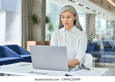 Serious Focused Asian Older 40s Businesswoman Executive Manager Sitting At Desk Working Typing On Pc Laptop Computer In Contemporary Office. Business Online Technology Concept.
