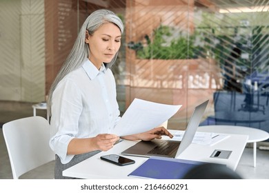 Serious Focused Asian Older Business Woman Ceo Manager Sitting At Desk Doing Financial Paperwork Using Pc Laptop Making Online Tax, Loans, Debts Bank Payments In Contemporary Corporation Office.