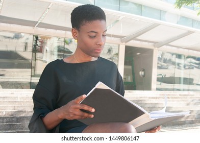 Serious Focused Black Woman Sitting On Stairs Outside And Studying Photo Book. African American Lady Reading Book Or Viewing Folder With Documents. Reader Concept