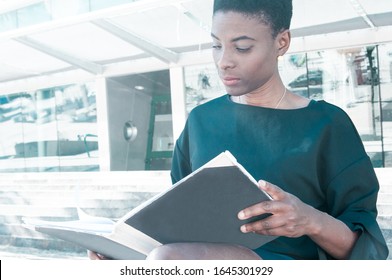 Serious Focused Black Woman Sitting On Stairs Outside And Studying Photo Book. African American Lady Reading Book Or Viewing Folder With Documents. Reader Concept