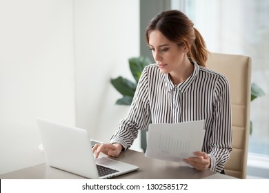 Serious focused businesswoman typing on laptop holding papers preparing report analyzing work results, female executive doing paperwork at workplace using computer online software for data analysis