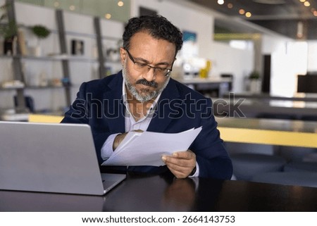 Serious focused elder Indian executive man reading document carefully, analyzing paper report at workplace with laptop, checking agreement text, bill, invoice for online payment