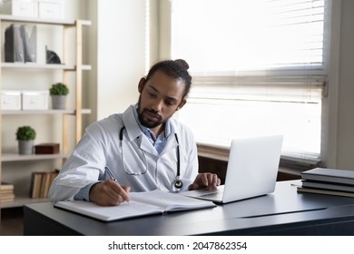 Serious focused GP doctor writing medical records at open laptop. Young Black practitioner listening to patient complaints, watching virtual medical training webinar, giving online video consultation