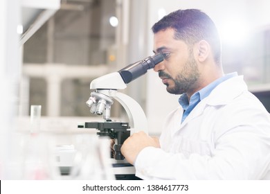 Serious Focused Handsome Young Arabian Biologist Sitting At Desk And Using Microscope While Analyzing Sample Of Modified Gene