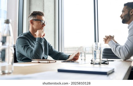 Serious focused mature businessman employer hr or client holding cv or contract listening manager, applicant during job interview, two diverse ethnic partners having negotiations at office meeting.